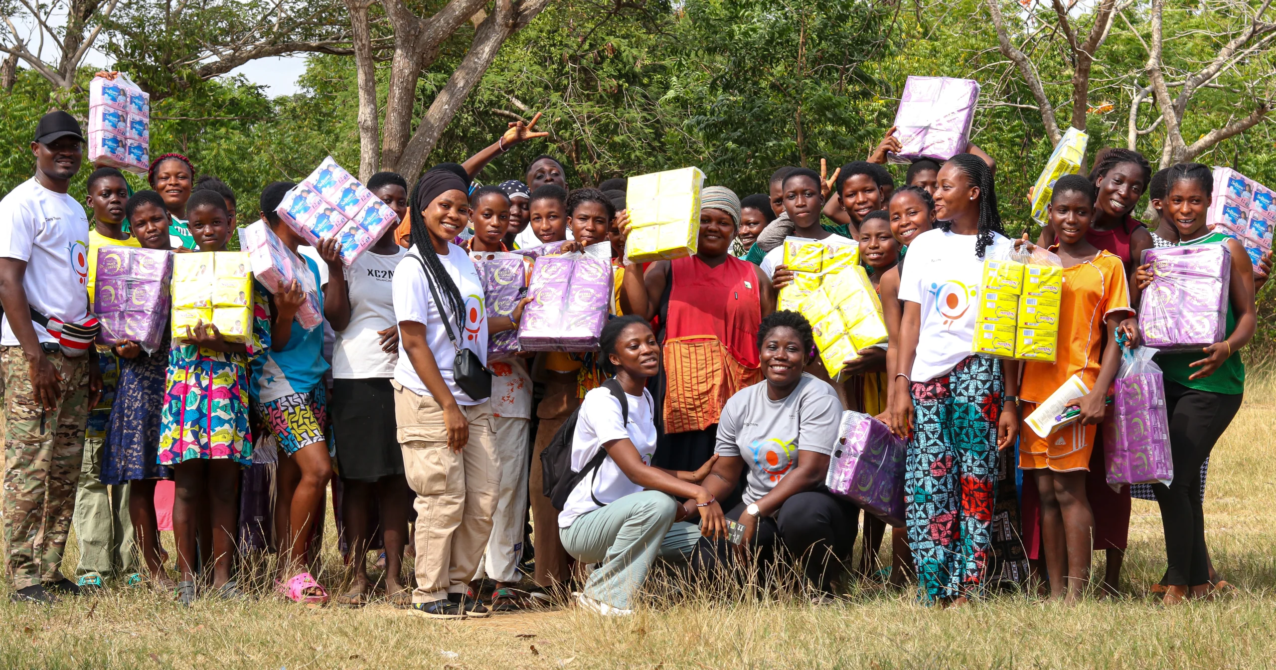 A group of young girls and volunteers smiling and holding up donated packages of sanitary pads during a Menstrual Hygiene Day community outreach event.