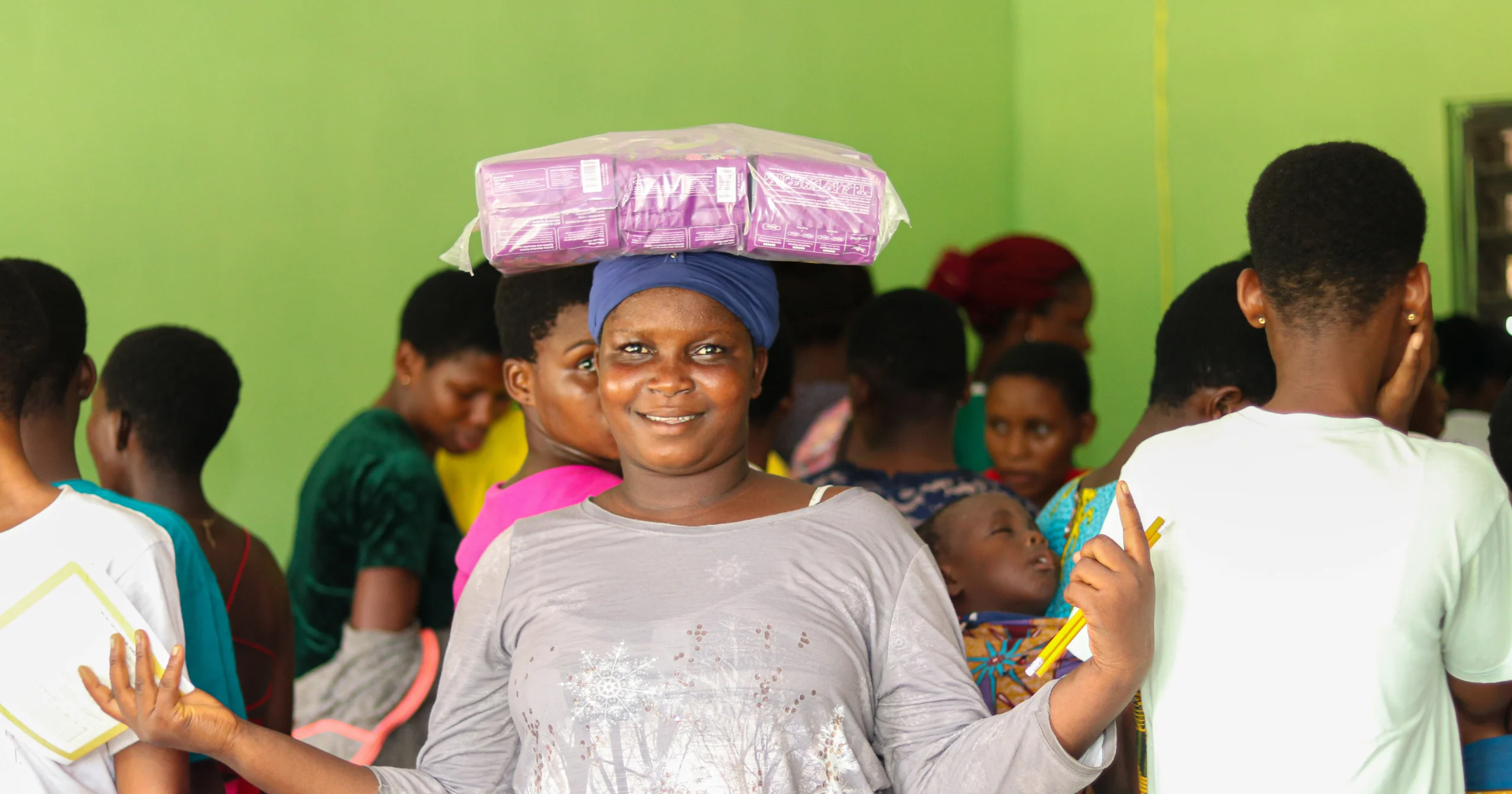 A smiling woman balancing a large pack of donated sanitary pads on her head during a Menstrual Hygiene Day community distribution event.