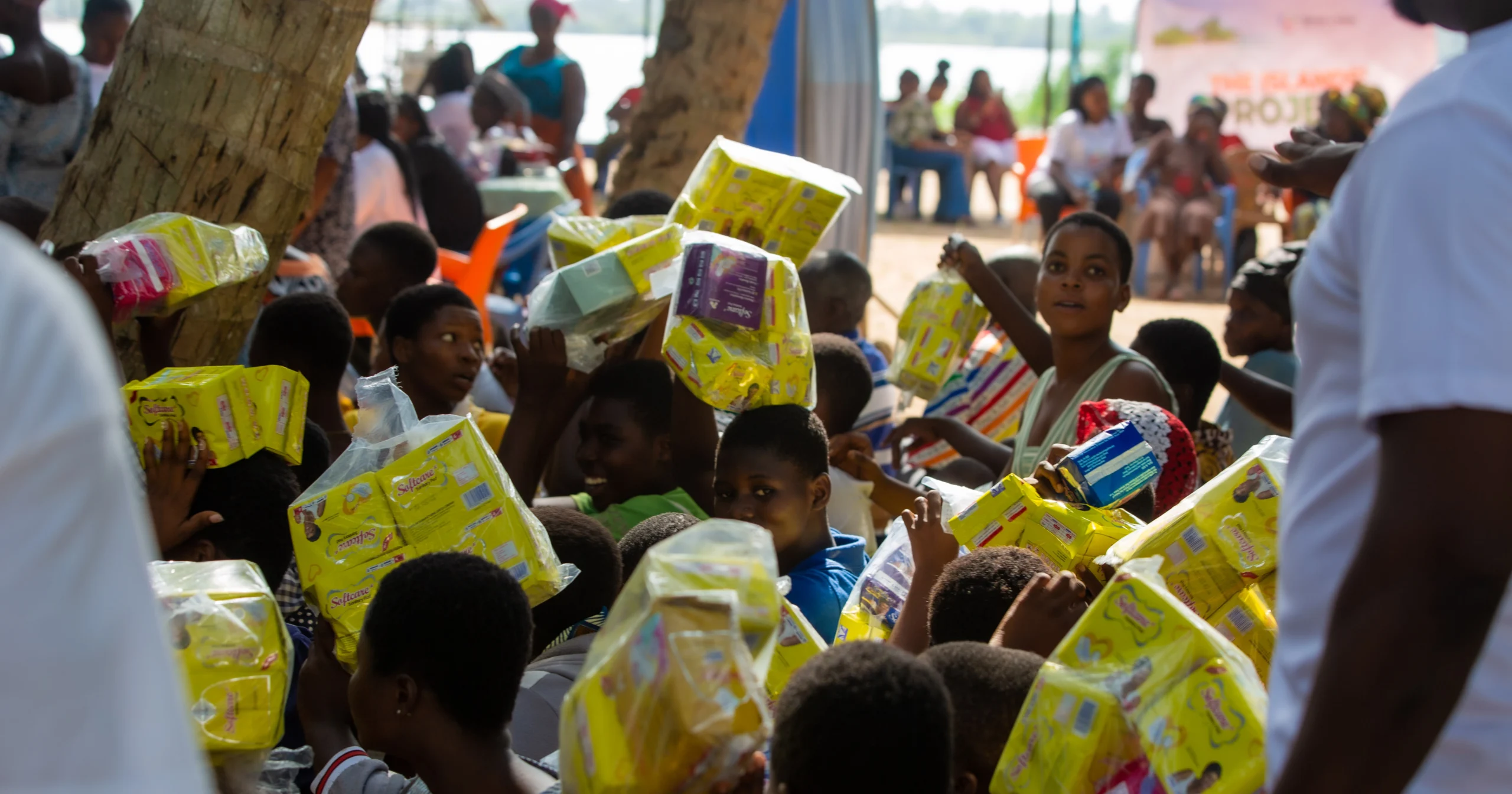 A crowd of young community members and children happily lifting up packages of donated sanitary pads during a Menstrual Hygiene Day outreach event for The Islands Project.