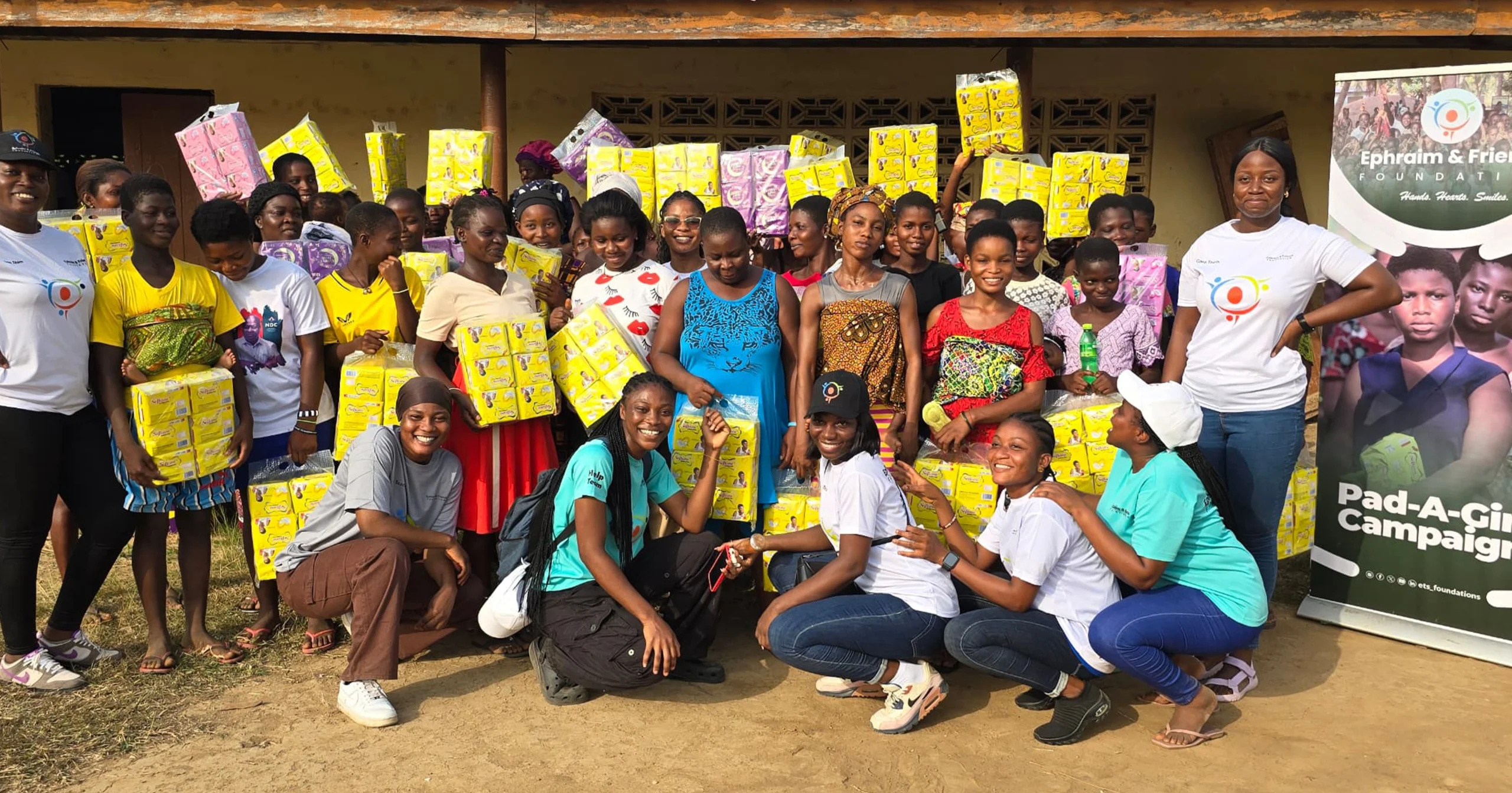 Volunteers and beneficiaries posing together with donated sanitary products next to an Ephraim & Friends Foundation Pad-A-Girl Campaign banner during a Menstrual Hygiene Day outreach event.
